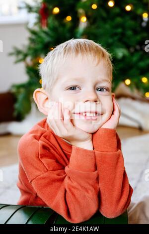 Portrait en gros plan d'un garçon mignon avec les mains sur le menton assis Avec arbre de Noël en arrière-plan Banque D'Images