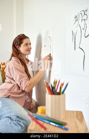 Artiste souriante dessinant avec du charbon de bois sur du papier collé mur dans la salle de séjour Banque D'Images
