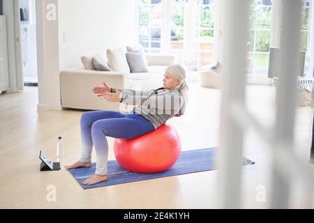 Femme senior s'exerçant sur le ballon de fitness tout en apprenant en ligne didacticiel sur la tablette numérique à la maison