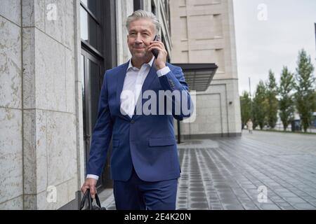 Homme d'affaires senior parlant sur téléphone portable tout en marchant bâtiment en ville Banque D'Images