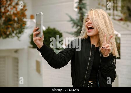 Jeune femme blonde faisant des gestes de paix tout en prenant le selfie à travers téléphone portable Banque D'Images