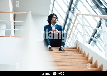 Homme d'affaires réfléchi regardant loin en étant assis avec les mains clastées sur l'escalier dans le bureau Banque D'Images