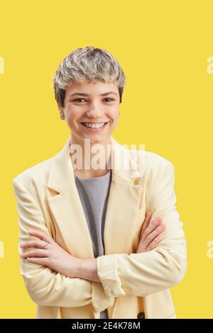 Portrait vertical à la taille haute d'une femme aux cheveux courts et confiante appareil photo debout, bras croisés sur fond jaune vif Banque D'Images