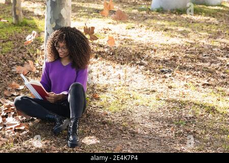 Femme souriante à la mode lisant le livre tout en se penchant près du tronc d'arbre au parc public Banque D'Images