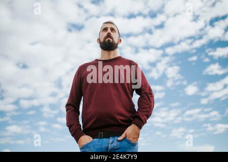 Homme barbu réfléchi avec les mains dans les poches debout contre le ciel nuageux ciel Banque D'Images