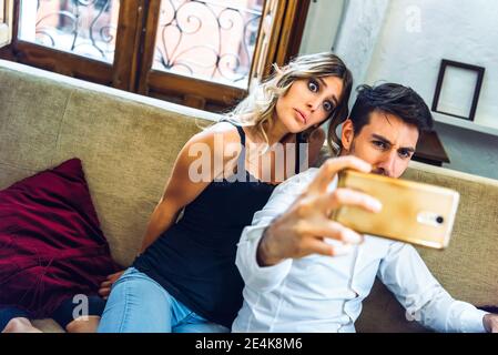 Un jeune couple fait des visages tout en prenant un selfie sur un canapé salle de séjour à la maison Banque D'Images