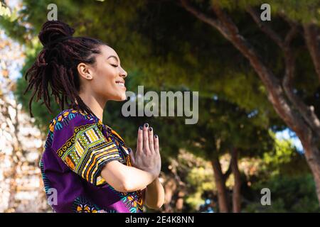 Souriante belle jeune femme méditant avec les mains jointes en pratiquant yoga au parc Banque D'Images