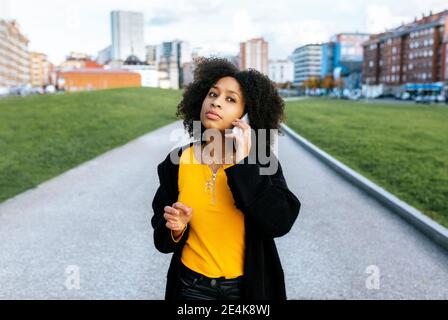 Jeune femme avec des cheveux afro parlant sur le smartphone pendant debout sur le sentier de la ville Banque D'Images