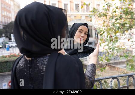 Jeune belle femme portant hijab noir debout à l'extérieur et regardant miroir rond Banque D'Images