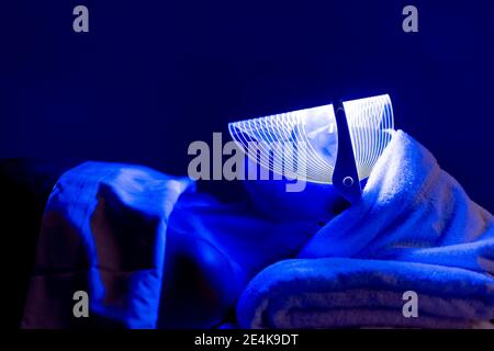 Jeune femme portant un masque bleu lors d'un traitement rajeunissant dans l'obscurité au centre de beauté Banque D'Images