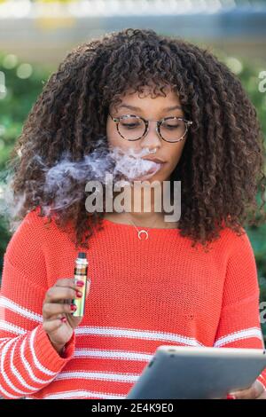 Femme afro portant des lunettes fumant la cigarette électronique tout en utilisant le numérique tablette au parc Banque D'Images