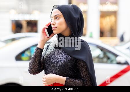 Portrait d'une jeune belle femme portant un hijab noir parlant sur un smartphone au milieu de la rue Banque D'Images