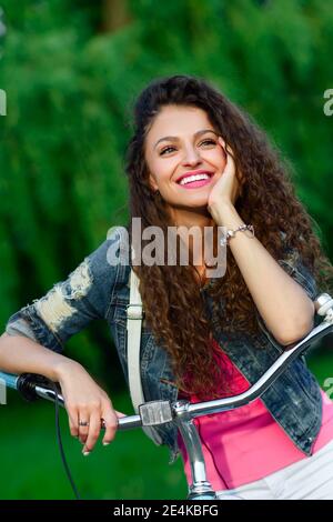 Belle fille aux cheveux bouclés sur un vélo dans le parc en été dans la ville. Portrait en extérieur Banque D'Images