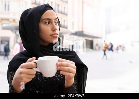 Portrait de jeune belle femme portant hijab noir appréciant une tasse de café au café-terrasse Banque D'Images