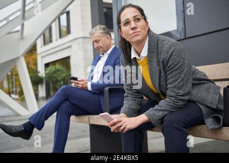 Navetteurs hommes et femmes avec smartphones assis sur un banc en attendant à l'arrêt de bus Banque D'Images