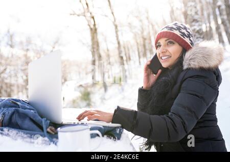 Femme d'affaires souriante avec un ordinateur portable parlant sur un téléphone intelligent pendant qu'elle est assise sur banc dans la neige Banque D'Images