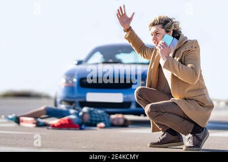 Jeune homme appelant de l'aide sur un téléphone mobile avec un garçon dedans arrière-plan allongé sur la route après un accident de voiture Banque D'Images