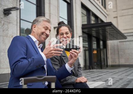 Des entrepreneurs hommes et femmes gais partageant un smartphone lorsqu'ils sont assis contre le bâtiment Banque D'Images