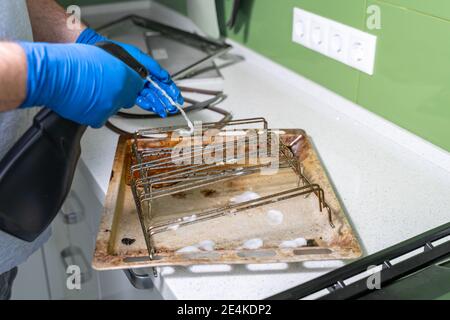 un homme portant des gants en caoutchouc bleu nettoie les grilles sales et plaques de cuisson du four avec un spray nettoyant Banque D'Images