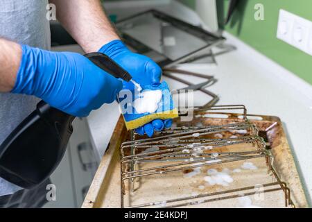 un homme portant des gants en caoutchouc bleu nettoie les grilles sales et plaques de cuisson du four avec un spray nettoyant Banque D'Images