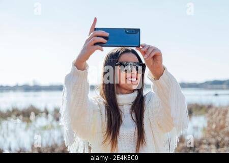 Belle femme portant des lunettes de soleil prenant selfie avec smartphone contre ciel dégagé Banque D'Images