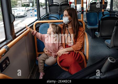 Mère avec une jolie fille pointant vers la fenêtre pendant son voyage Bus pendant COVID-19 Banque D'Images