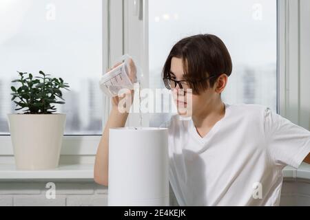 un jeune homme en verre et un t-shirt blanc verse de l'eau dans le réservoir d'un humidificateur. un homme utilise un humidificateur à la maison Banque D'Images