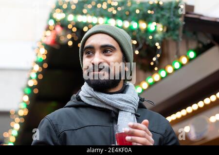 Gros plan sur un homme barbu bien pensé qui tient une boisson contre un Noël illuminé feux Banque D'Images