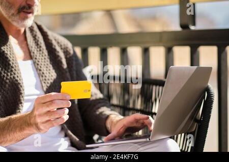 Homme mature à la maison avec carte de crédit faisant des achats en ligne sur ordinateur portable pendant la journée ensoleillée Banque D'Images