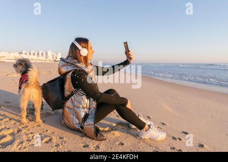 Femme prenant le selfie tout en étant assise avec un chien à la plage contre ciel dégagé au coucher du soleil Banque D'Images