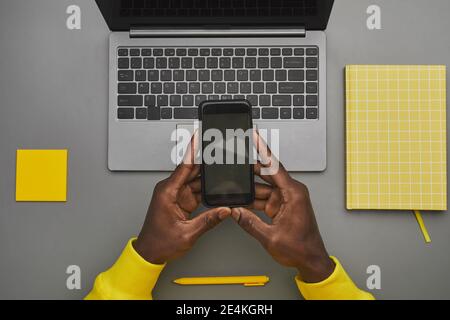 Arrière-plan gris et jaune représentant des mains afro-américaines de sexe masculin tenant un smartphone avec un écran vide sur un ordinateur portable tout en travaillant à un bureau, de haut en bas vie Banque D'Images