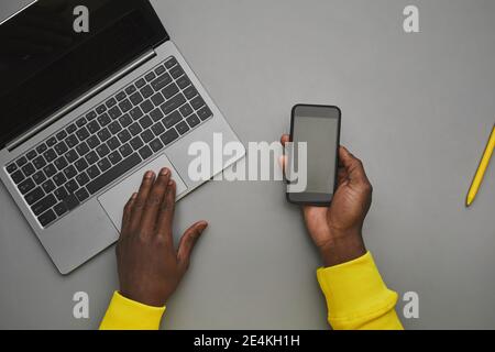 Arrière-plan gris minimal de mains afro-américaines tenant un smartphone avec écran vide au-dessus d'un ordinateur portable pendant que vous travaillez au bureau, vue du haut vers le bas, Copy spa Banque D'Images