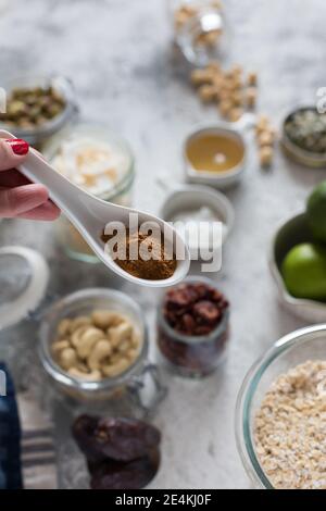 Une femme tient à la main une cuillère en céramique remplie de cannelle en poudre sur quelques ingrédients de granola flous. Banque D'Images