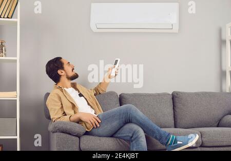 Jeune homme assis sur un canapé à la maison et allumé climatiseur avec télécommande Banque D'Images