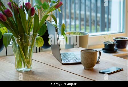 Bureau à domicile moderne au printemps. Ordinateur portable, téléphone portable, tasse à café et tulipes fraîches dans un vase sur une table en bois. Photo prise en Suède. Banque D'Images