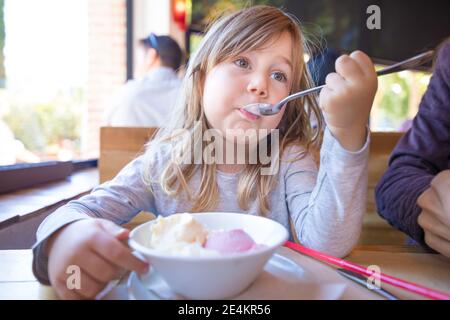 portrait d'un enfant blond de trois ans qui mange de la glace à la fraise et à la vanille avec une cuillère dans un bol blanc, assis à la table du restaurant Banque D'Images