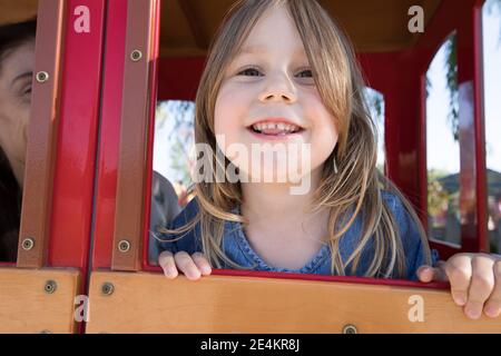 face de la jeune fille blonde de quatre ans penchée par la fenêtre de la maison de jeux dans le parc public, à côté de la mère Banque D'Images