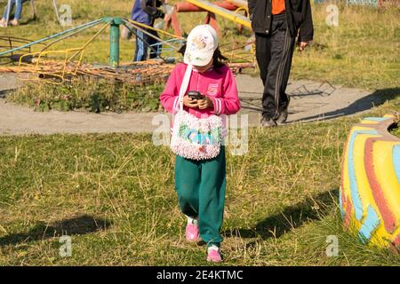 Une petite fille avec un téléphone portable dans ses mains marche autour de la cour lors d'un jour d'automne ensoleillé. Banque D'Images