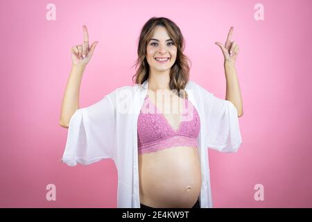 Jeune belle femme brune enceinte s'attendant à bébé sur fond rose isolé sourire, regardant l'appareil photo et pointant vers le haut avec les doigts et r Banque D'Images