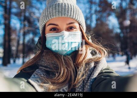 Femme portant un masque de protection dans un parc hivernal enneigé prenant le selfie. Mesures de sécurité en extérieur pour la pandémie de virus Corona Covid-19. Banque D'Images