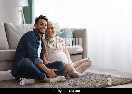 Heureux couple musulman enceinte se relaxant à la maison ensemble, assis à l'étage Banque D'Images