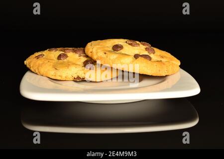 Deux biscuits entiers ronds, de couleur sable, parfumés et frais avec morceaux de chocolat au lait, sur une soucoupe blanche, sur fond noir. Banque D'Images