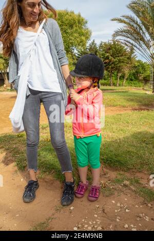 Blonde de quatre ans heureuse fille souriante, tenant la main de la mère avec un chapeau d'équitation, attendant de monter un cheval Banque D'Images