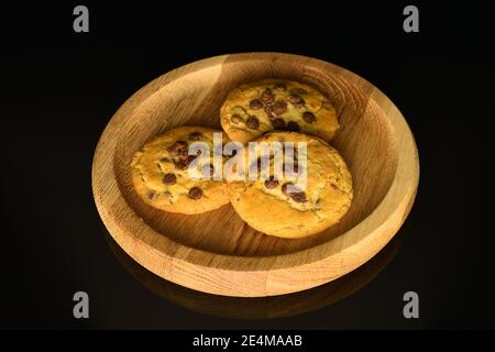 Sur un plateau rond en bois se trouvent trois biscuits sablés avec des morceaux de chocolat au lait. L'arrière-plan est noir. Banque D'Images