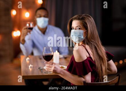 Date de la pandémie. Jeune couple interracial en masques médicaux dîner au restaurant Banque D'Images