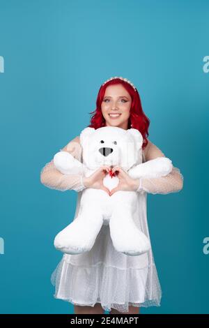 rire drôle femme dans une robe blanche avec des cheveux rouges épouse délicatement un ours en peluche blanc et fait un cœur de ses doigts isolés sur un fond bleu Banque D'Images