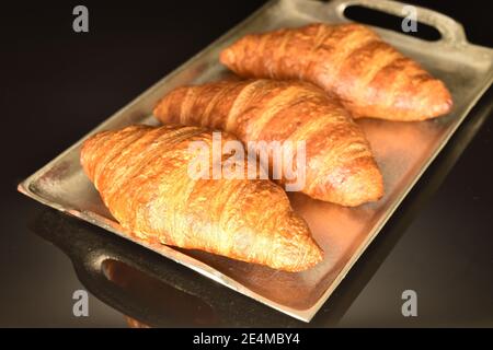 Sur fond noir, trois croissants croquants, frais et parfumés, se trouvent sur un plateau en métal Banque D'Images