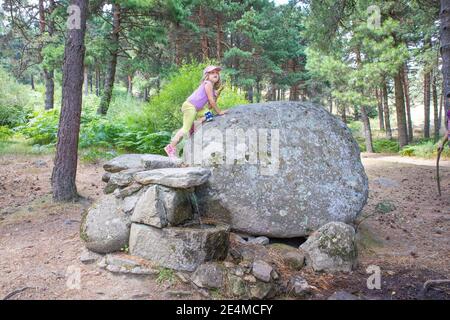 Jeune fille blonde de cinq ans grimpant sur un grand rocher, au-dessus d'une fontaine, en été, dans une forêt du parc naturel de Guadarrama (Madrid, Espagne, Europe) Banque D'Images