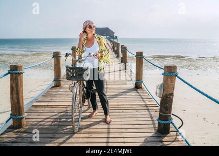 Portrait d'une femme souriante vêtue de vêtements d'été légers, lunettes de soleil avec vélo sur la jetée en bois de la mer sur la plage de sable de Zanzibar.négligent vacat Banque D'Images
