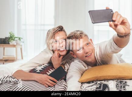 Jeunes adultes gaies Près en pyjama prendre une photo de selfie à l'aide d'un smartphone moderne comme paresseux reposant allongé dans un lit confortable dans la chambre. Banque D'Images